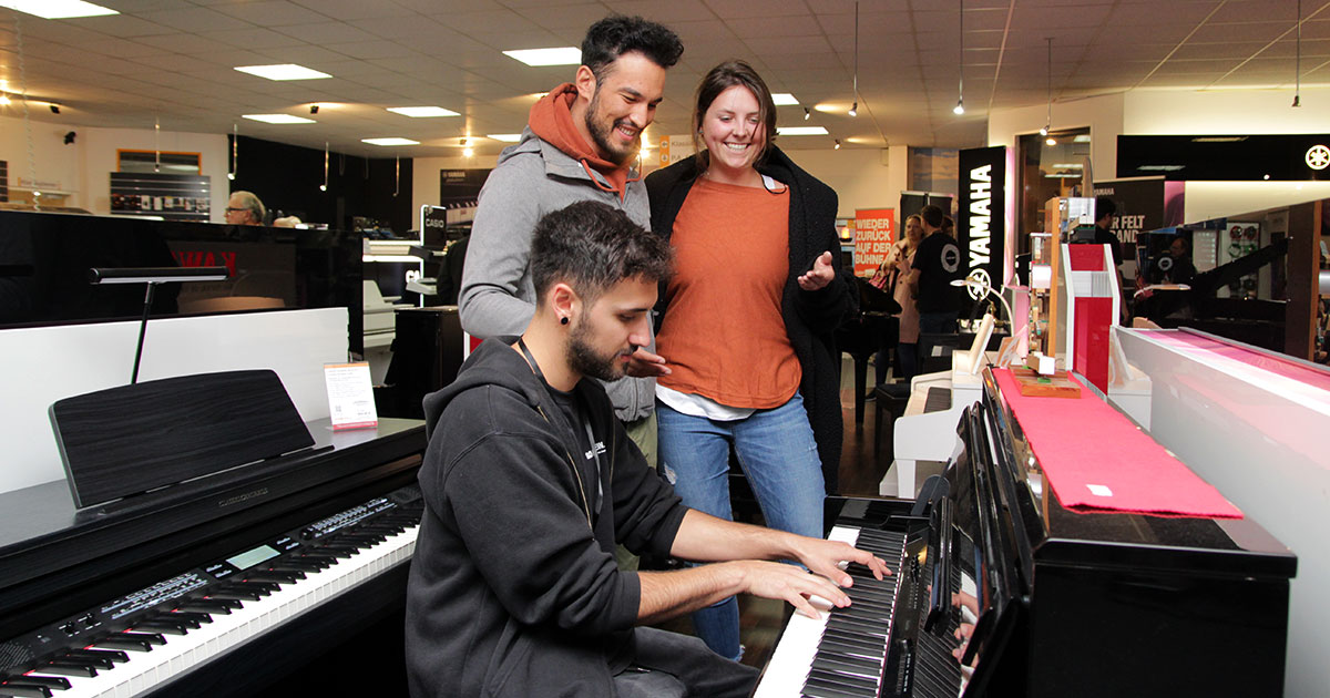 Besucher der MusiConnect 2022 schauen einem Kirstein-Mitarbeiter in der Digitalpiano-Abteilung beim Klavierspielen zu.