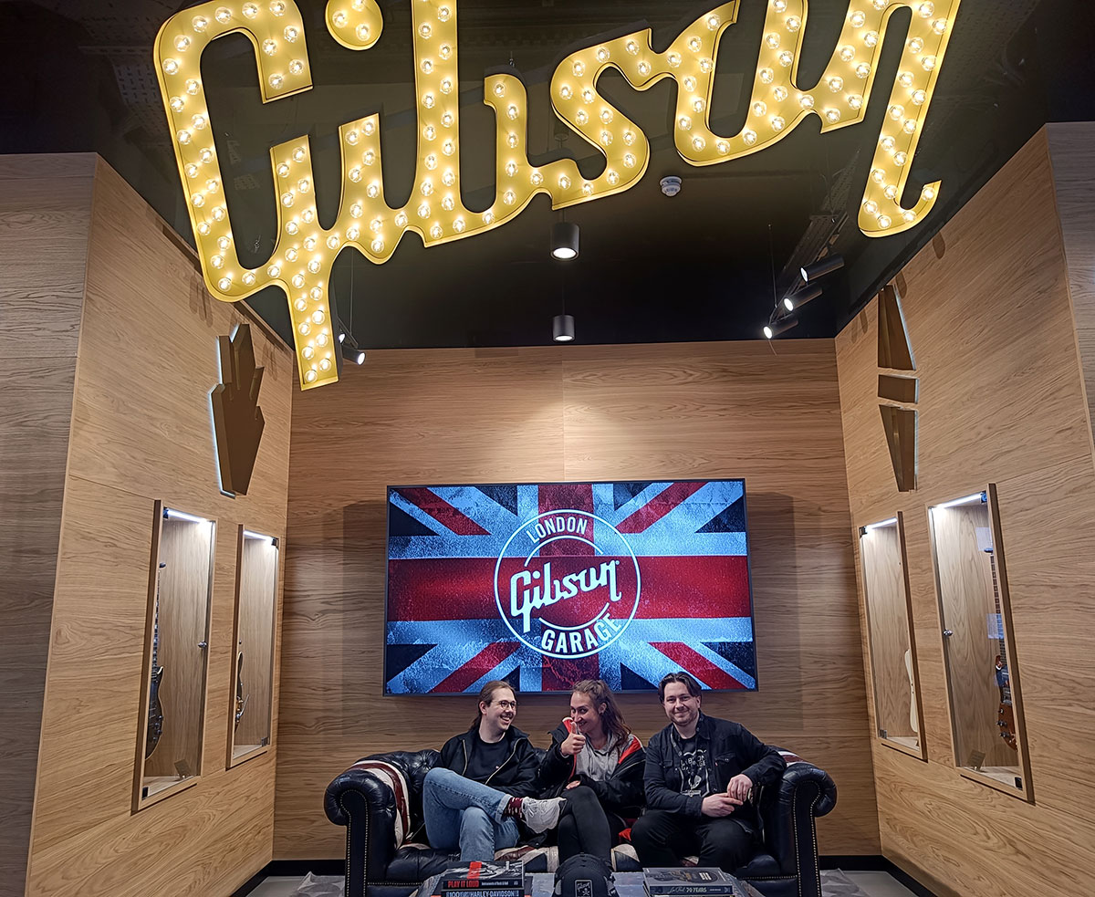 In der Gibson Garage sitzen Nico, Mali und Sam Gammon auf einem Ledersofa, im Hintergrund die Flagge Großbritanniens mit dem Gibson-Garage-Logo.