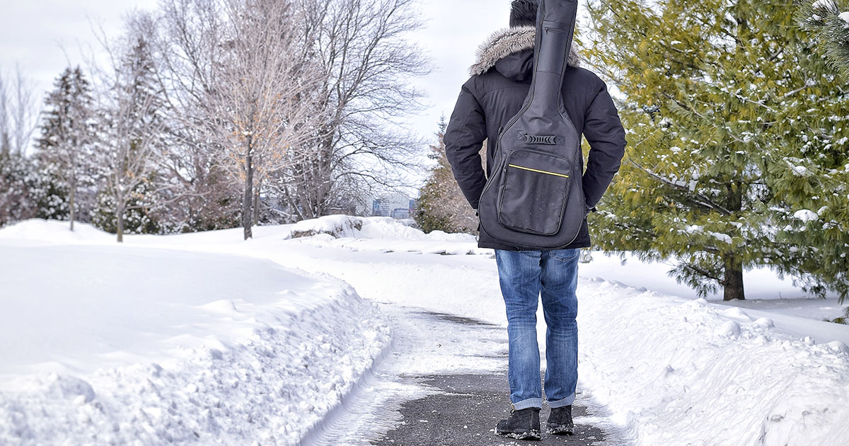 Mann, der in einer verschneiten Landschaft mit einer Gitarrengigbag auf dem Rücken zu Fuß unterwegs ist.